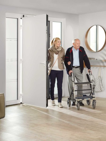 Frau begleitet älteren Mann mit einem Rollator ins Haus